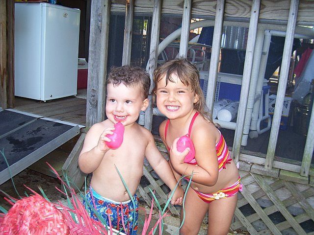 Two children smiling, holding pink objects, wearing swimsuits, on a wooden porch.
