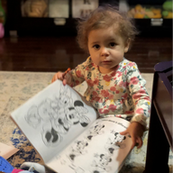 Child holding open coloring book, looking up with a neutral expression. Sitting on a rug.