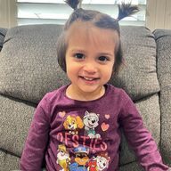 Young child with pigtails wearing a purple Paw Patrol shirt smiles while sitting on a couch.