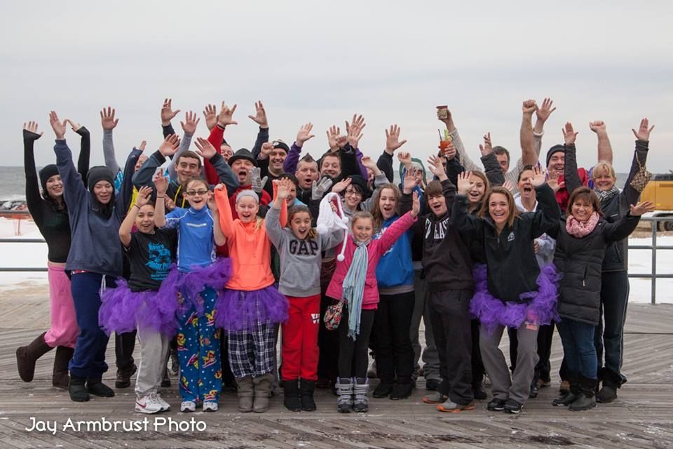 Group of people with arms raised, on a boardwalk, many wearing tutus.