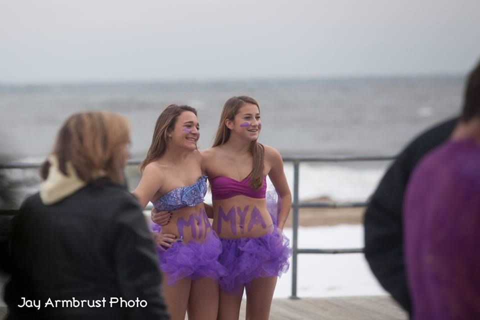 Two girls in purple tutus and decorated tops pose on a boardwalk near the ocean.