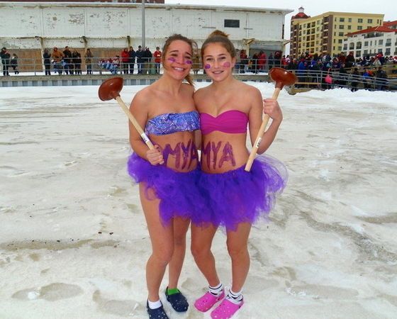 Two people in tutus and swimwear holding mallets, standing on ice. Purple and blue outfits, cheering crowd.