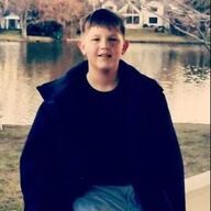Young person in a dark coat smiles, standing in front of a pond and houses.