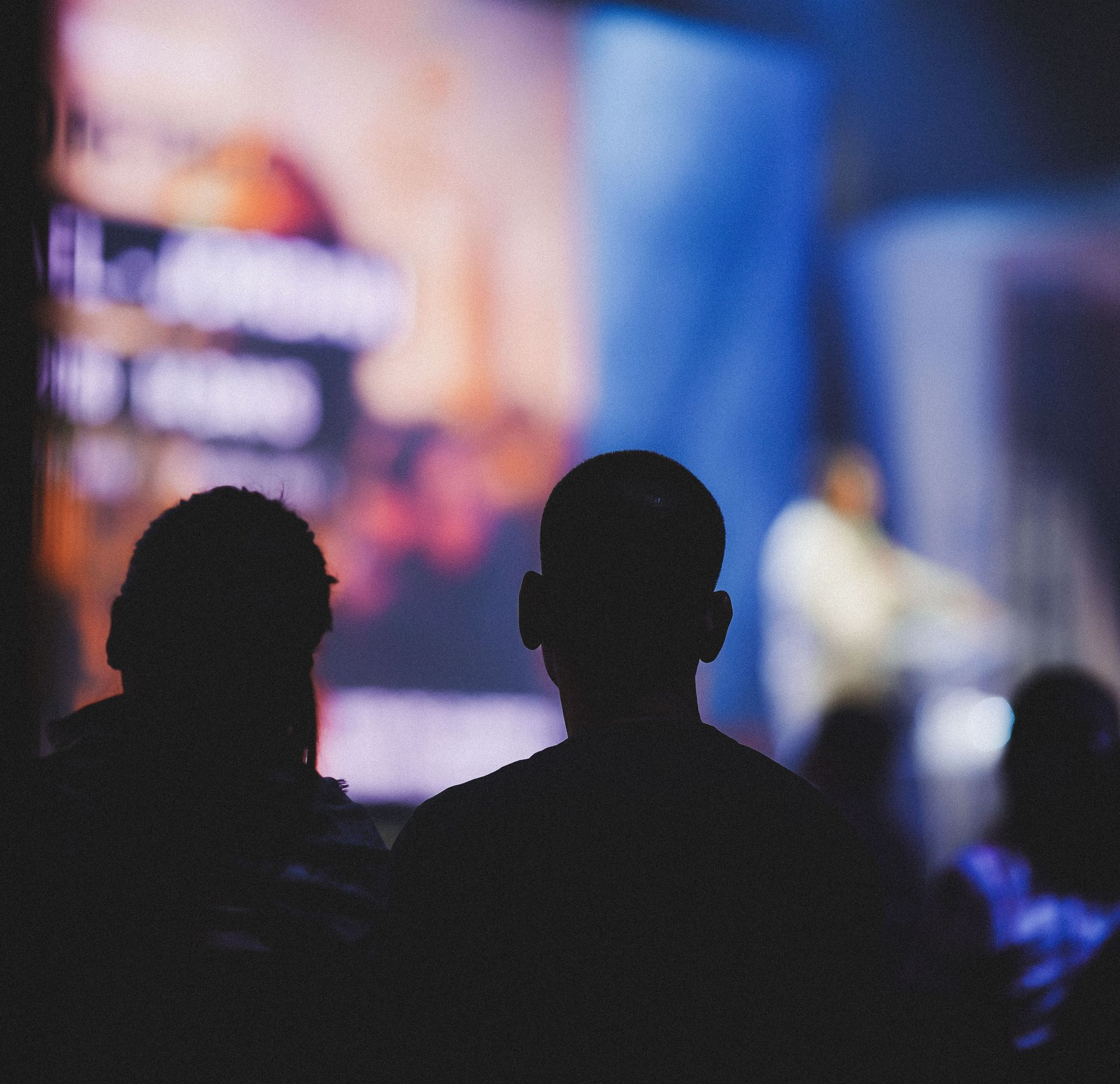 Two dark silhouettes seated before a blurry stage with blue and orange lights