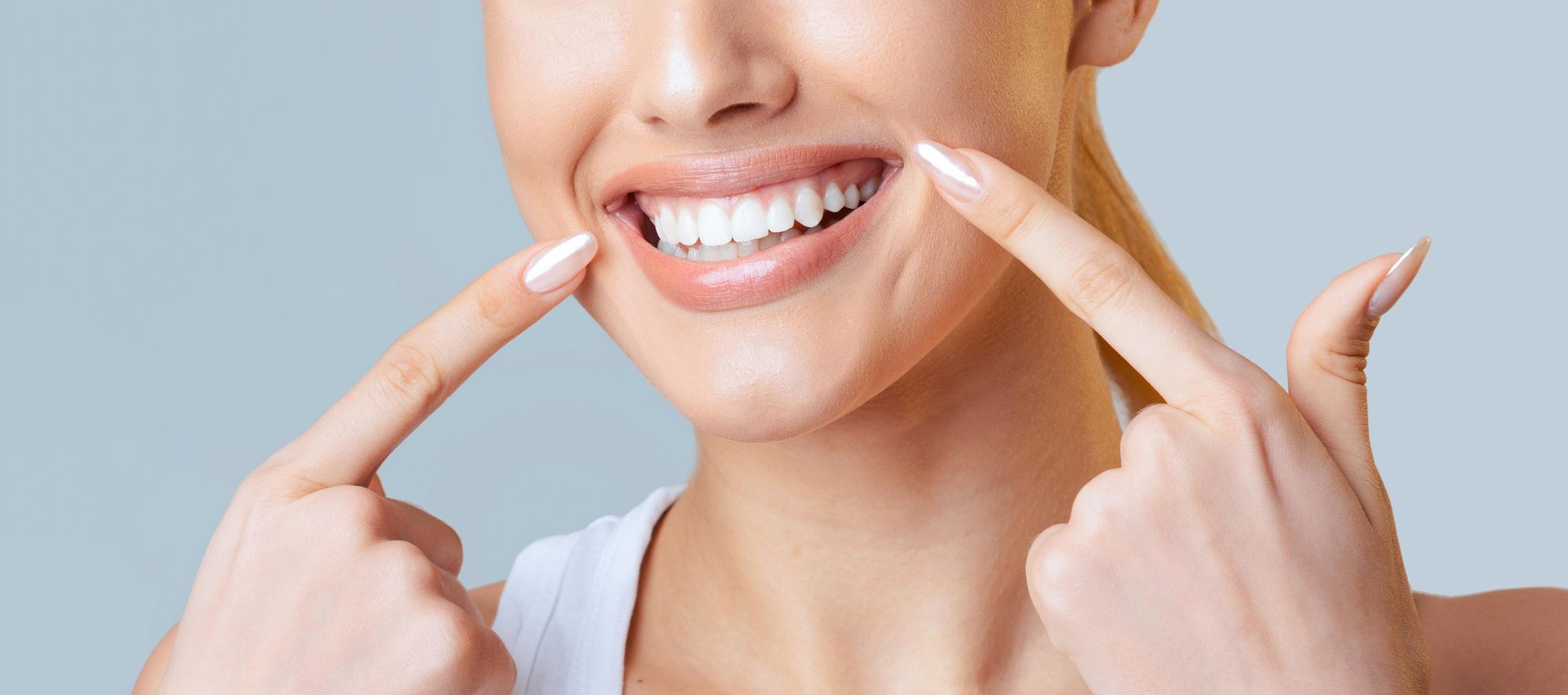 A person pointing to their straight, white teeth against a plain light-blue background.