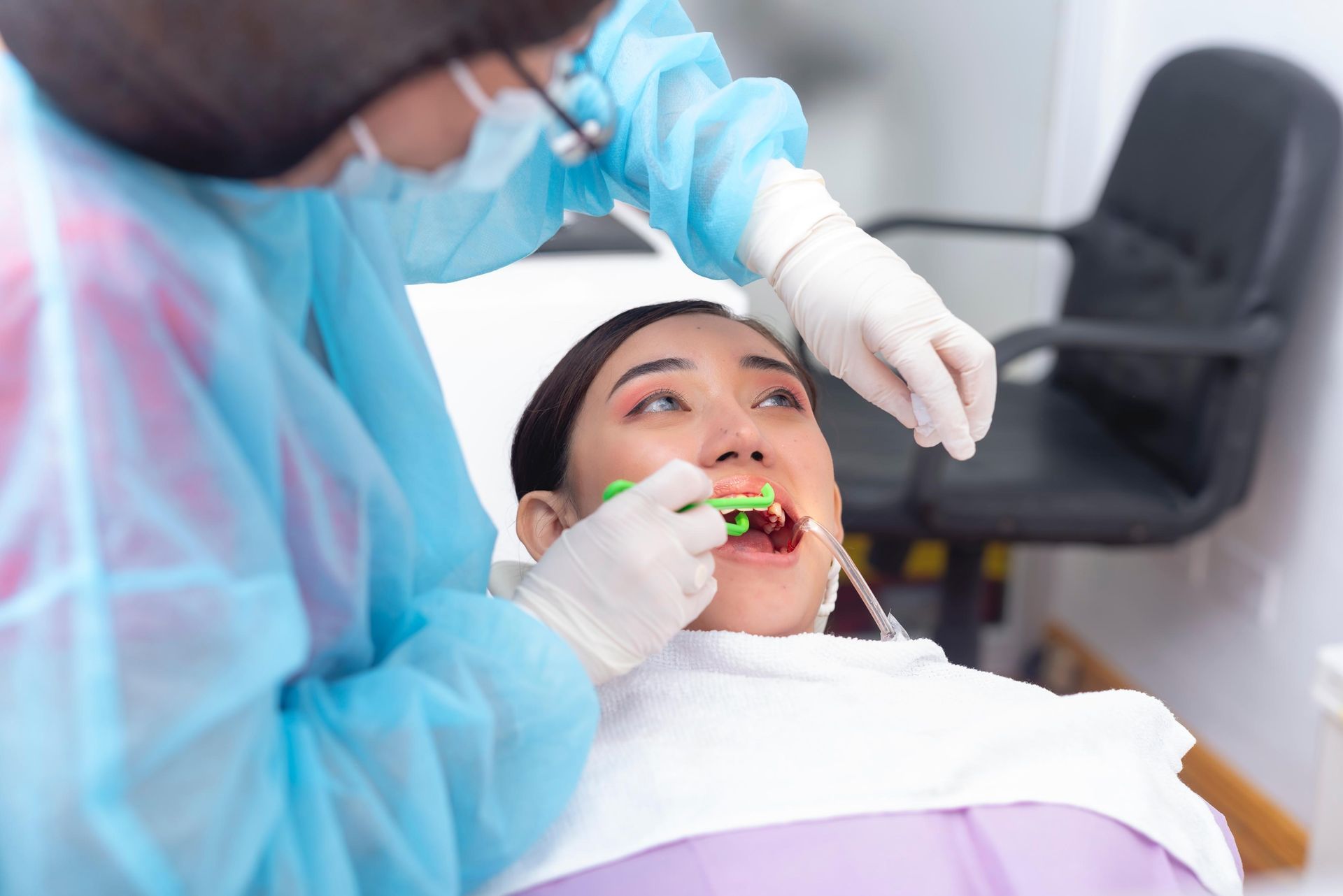 A dentist wearing blue scrubs and gloves performs a dental procedure on a patient in a clinic chair.