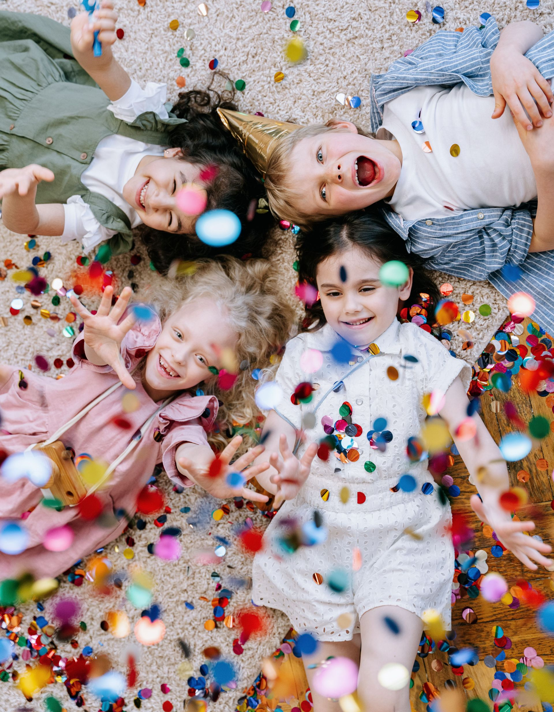 Four children lying on a confetti-covered floor, smiling with colorful candies scattered around them