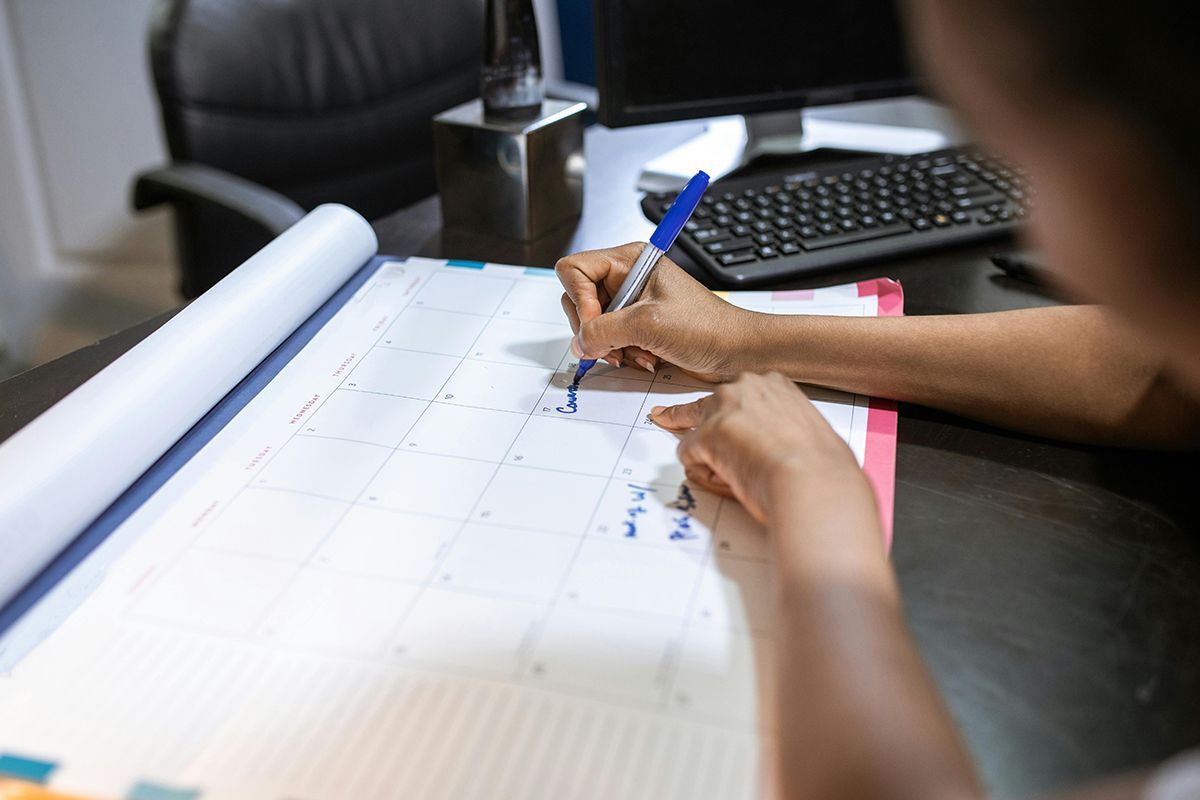 Hands drawing on a large calendar planner at a desk with a keyboard and chair nearby