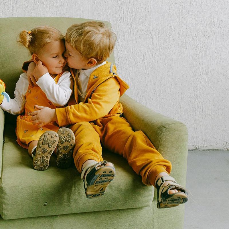 Two young children on a green couch, hugging and kissing, wearing mustard-colored clothes.