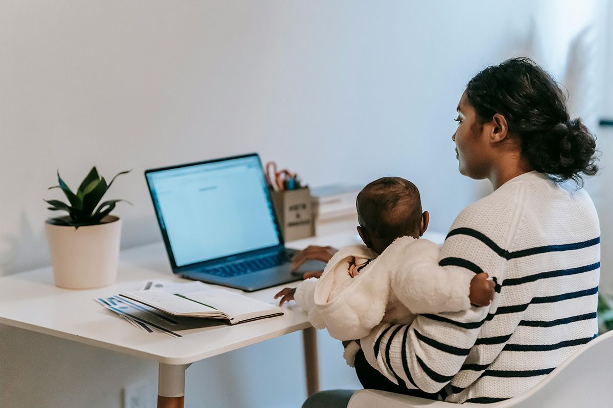 Woman holding a baby at a desk using a laptop in a bright home office