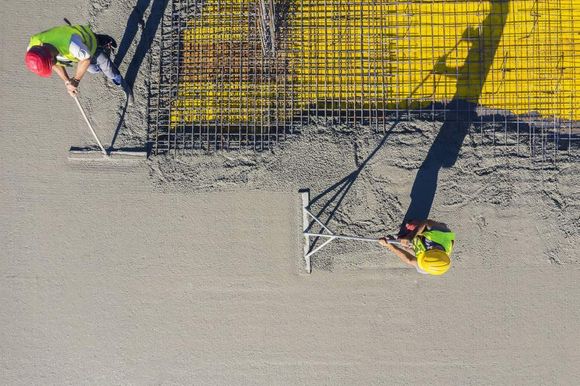 Operai edili spargono cemento fresco in un cantiere; un operaio indossa un casco rosso, un altro uno giallo.