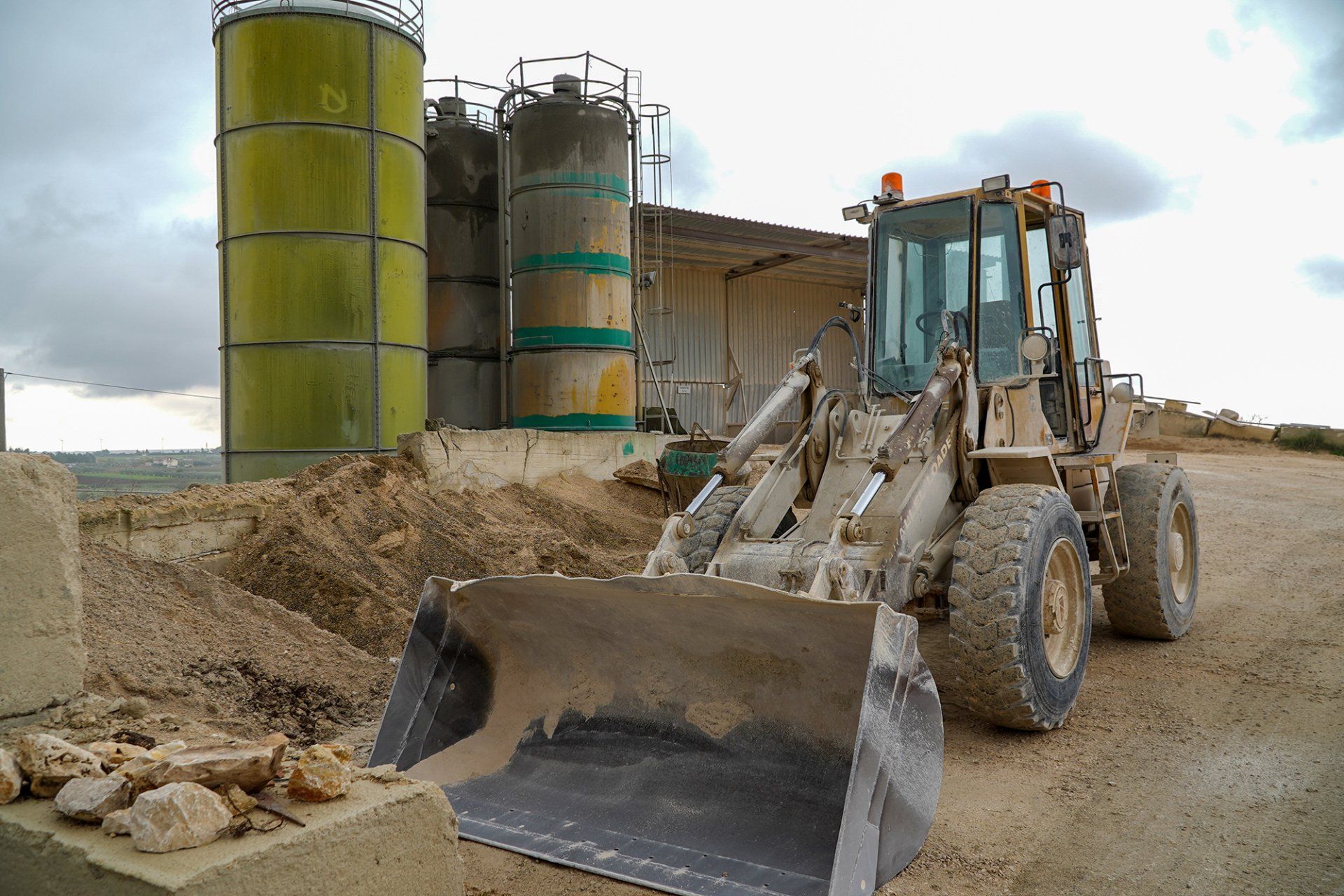 Pala caricatrice frontale in un cantiere edile vicino a serbatoi di stoccaggio gialli, che raccoglie detriti sotto un cielo nuvoloso.