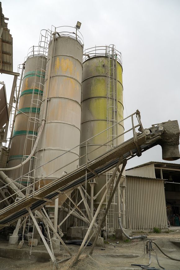 Tre grandi silos industriali, di diverse tonalità di grigio e verde, con scale e nastro trasportatore.