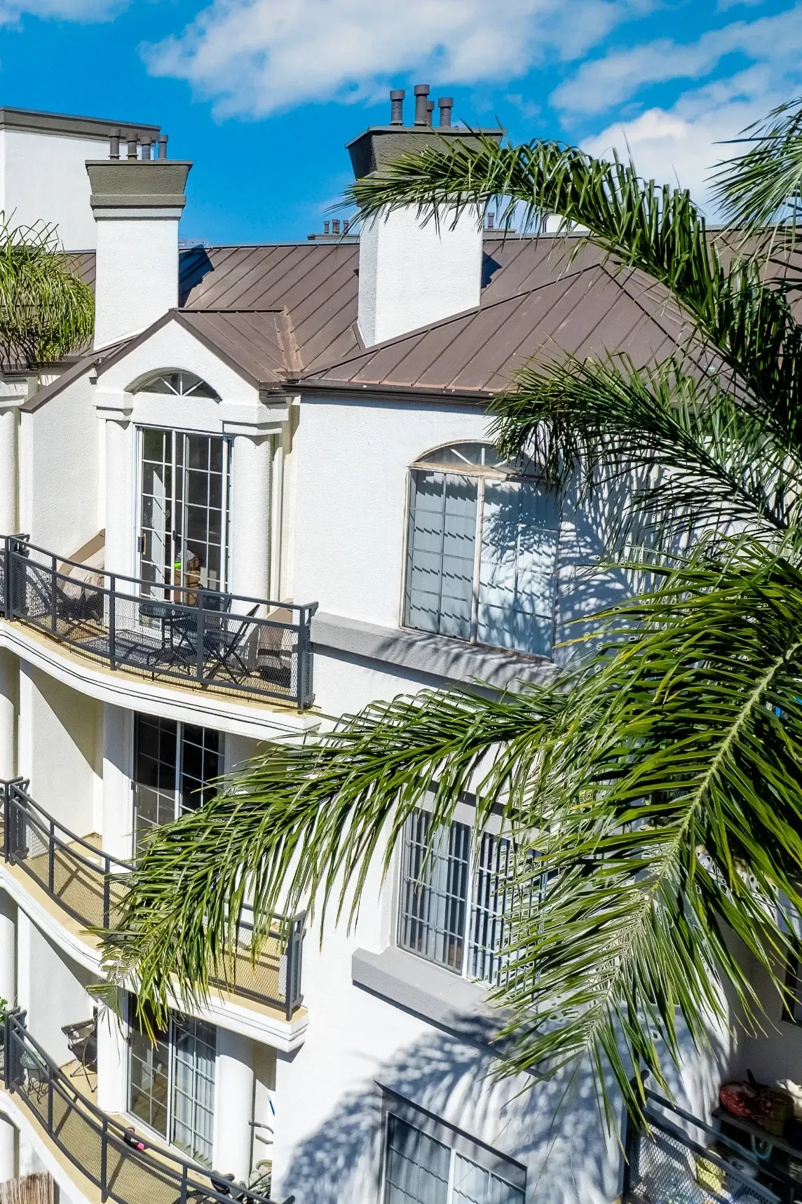 Exterior view of a multi-story apartment building with white stucco walls, balconies, and palm fronds in the foreground.