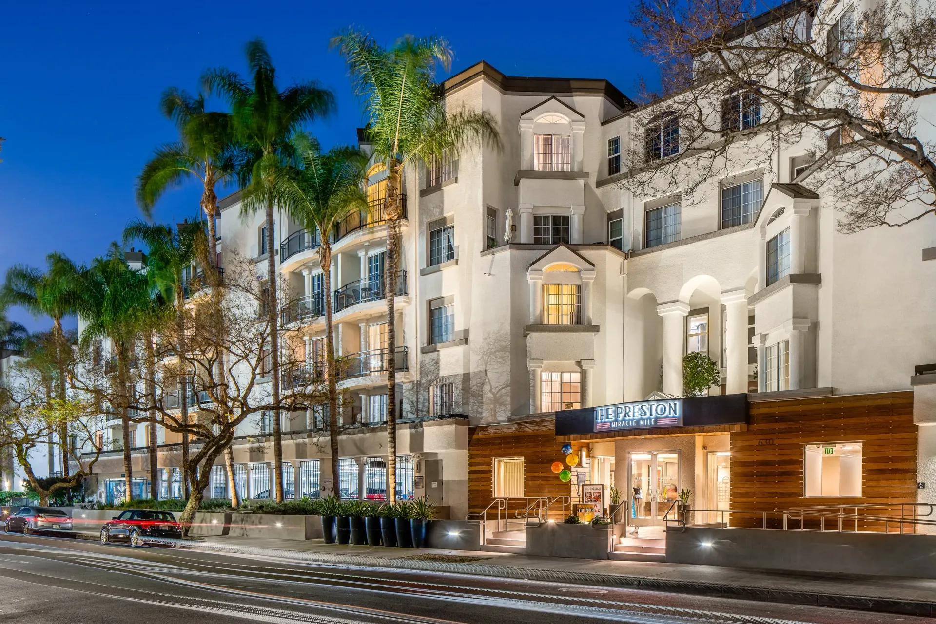 The Preston at Miracle Mile apartment building exterior at dusk with palm trees and streetlights.