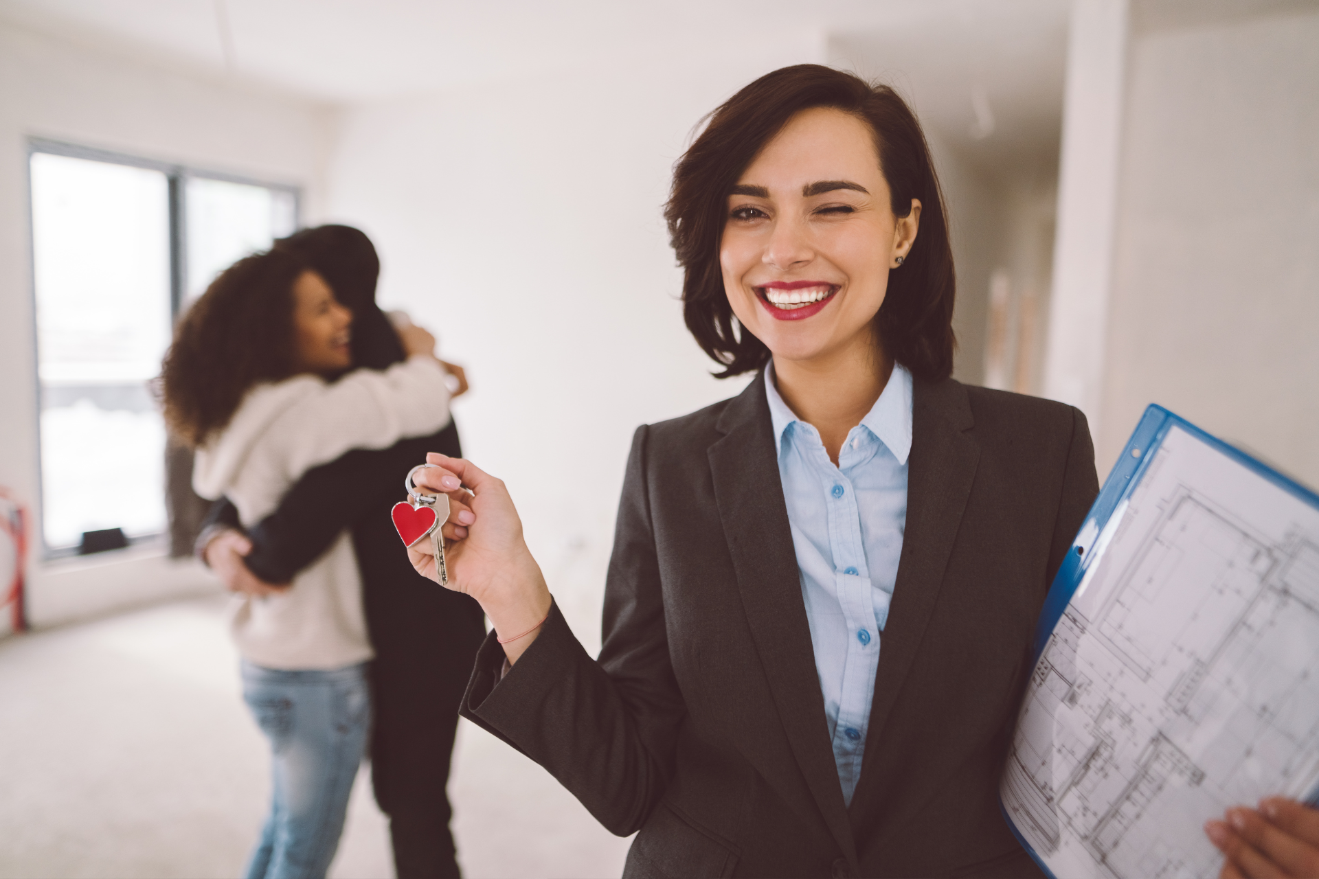 Smiling professional holding keys in a bright room, with another person hugging in the background and blueprints in hand