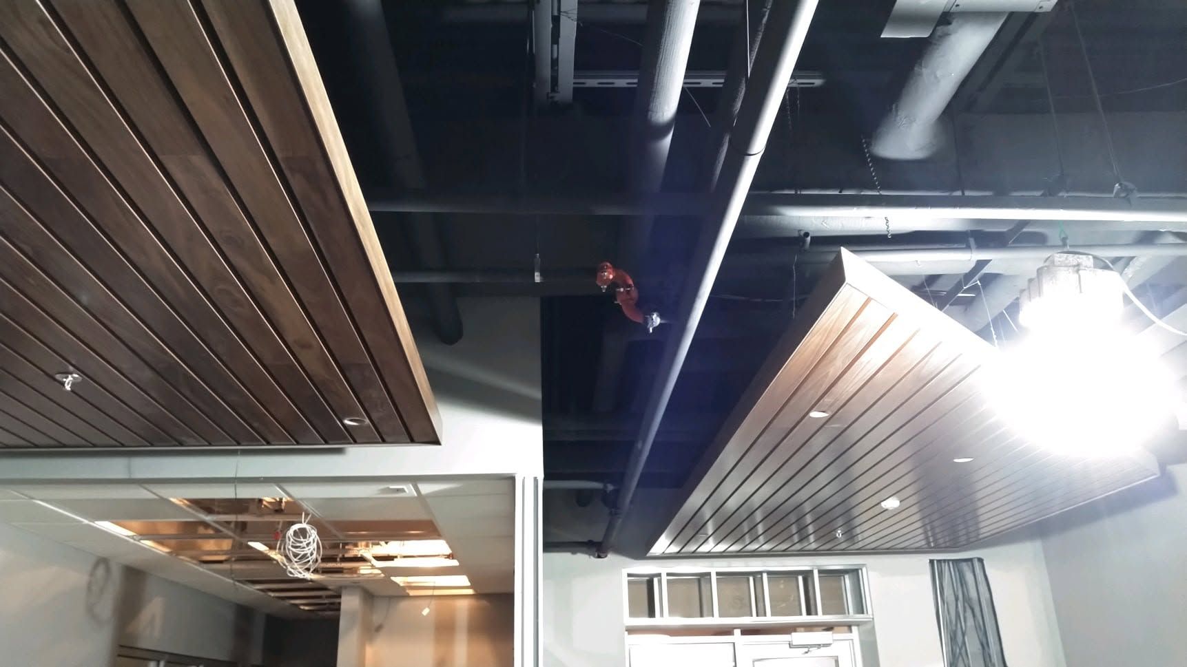 Modern indoor ceiling with wood slats, exposed pipes, and bright hanging lights in a room under construction