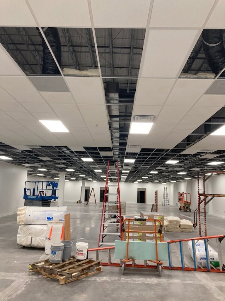 Office interior under renovation with exposed ceiling tiles, ladders, and construction materials scattered around.