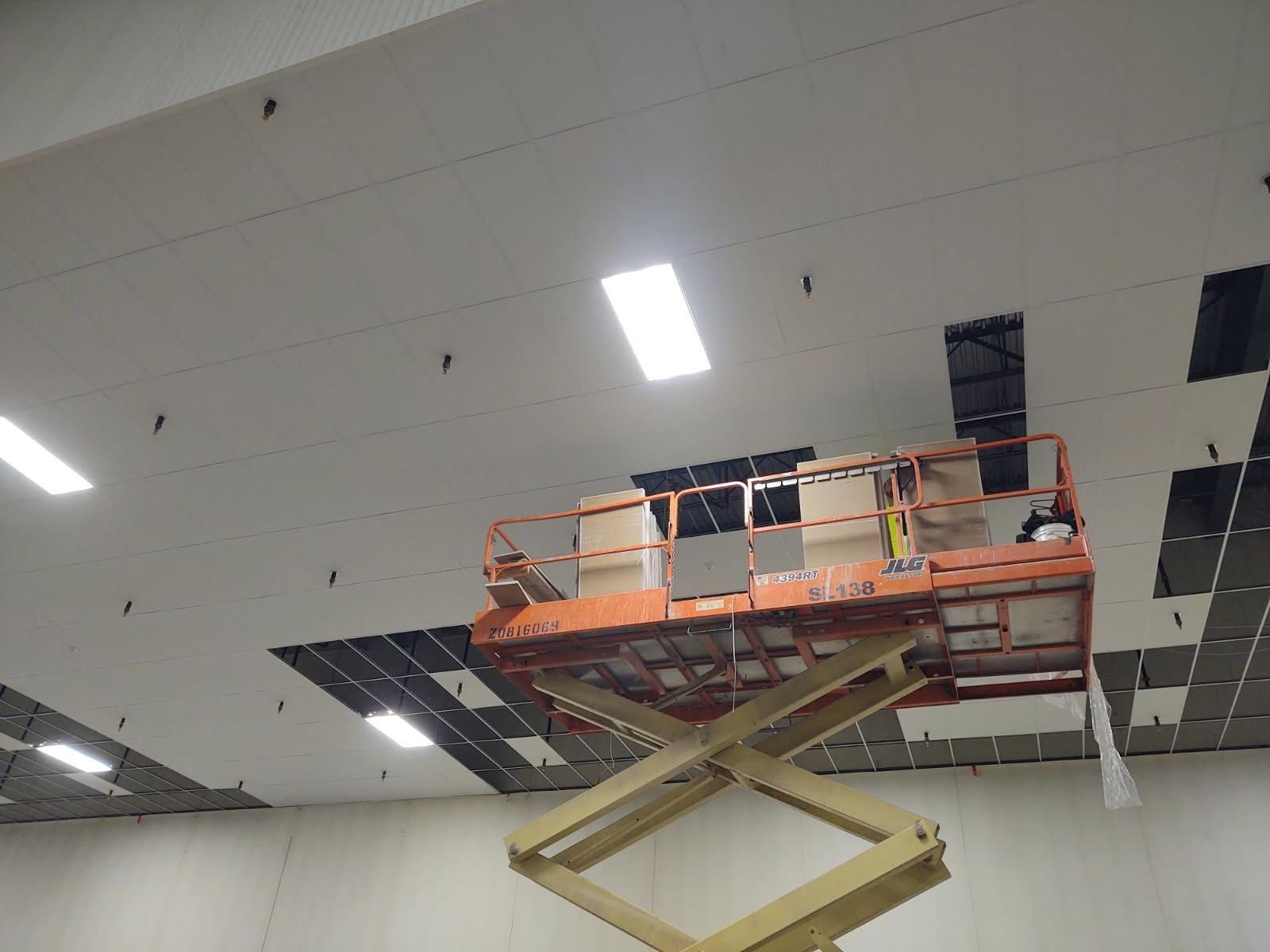 Orange scissor lift raising workers under a damaged ceiling with missing panels and exposed sections