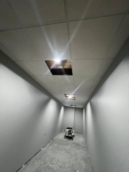 Narrow gray hallway under construction with exposed ceiling lights and a small cart on the floor.