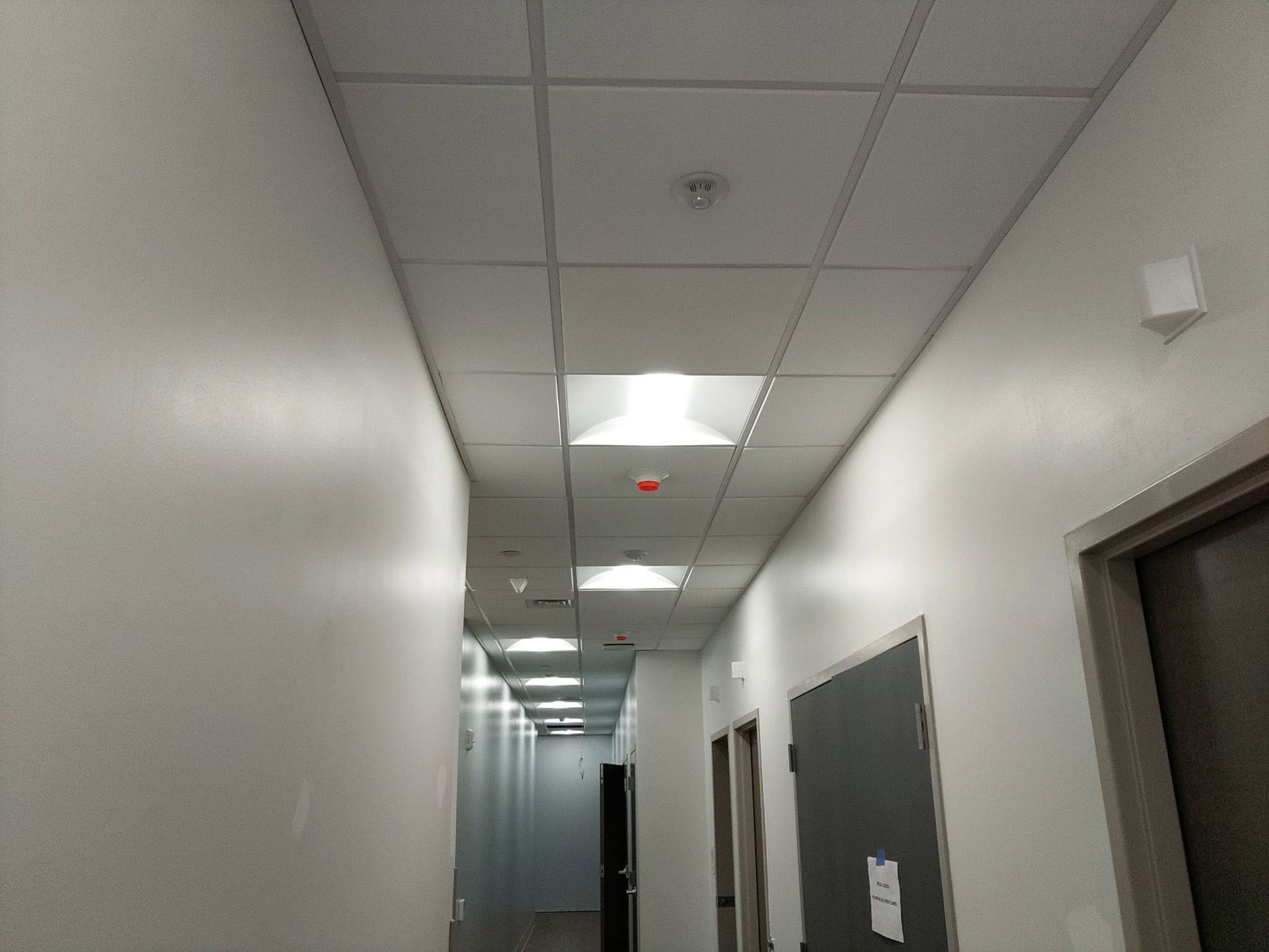 Narrow office hallway with white walls, gray floor, ceiling lights, and a dark open doorway on the right