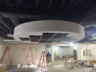 Construction site with circular white ceiling feature being installed beneath exposed ceiling framing.