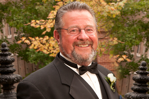 Smiling man in a black tuxedo with a white boutonniere, standing outdoors in front of autumn foliage.