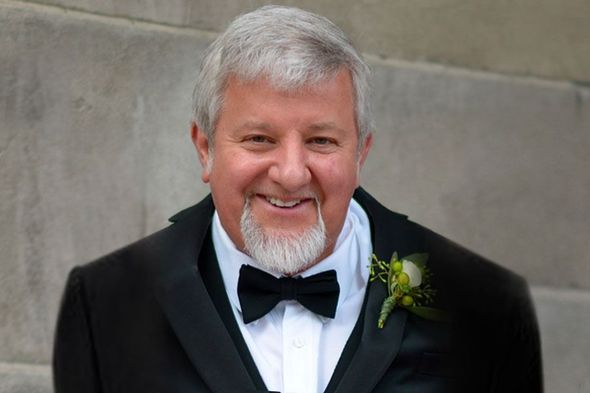 Smiling man in a black tuxedo with a bow tie and boutonniere against a gray wall