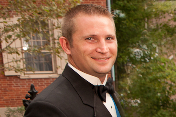 Man in a black tuxedo smiling outdoors in front of a building and trees