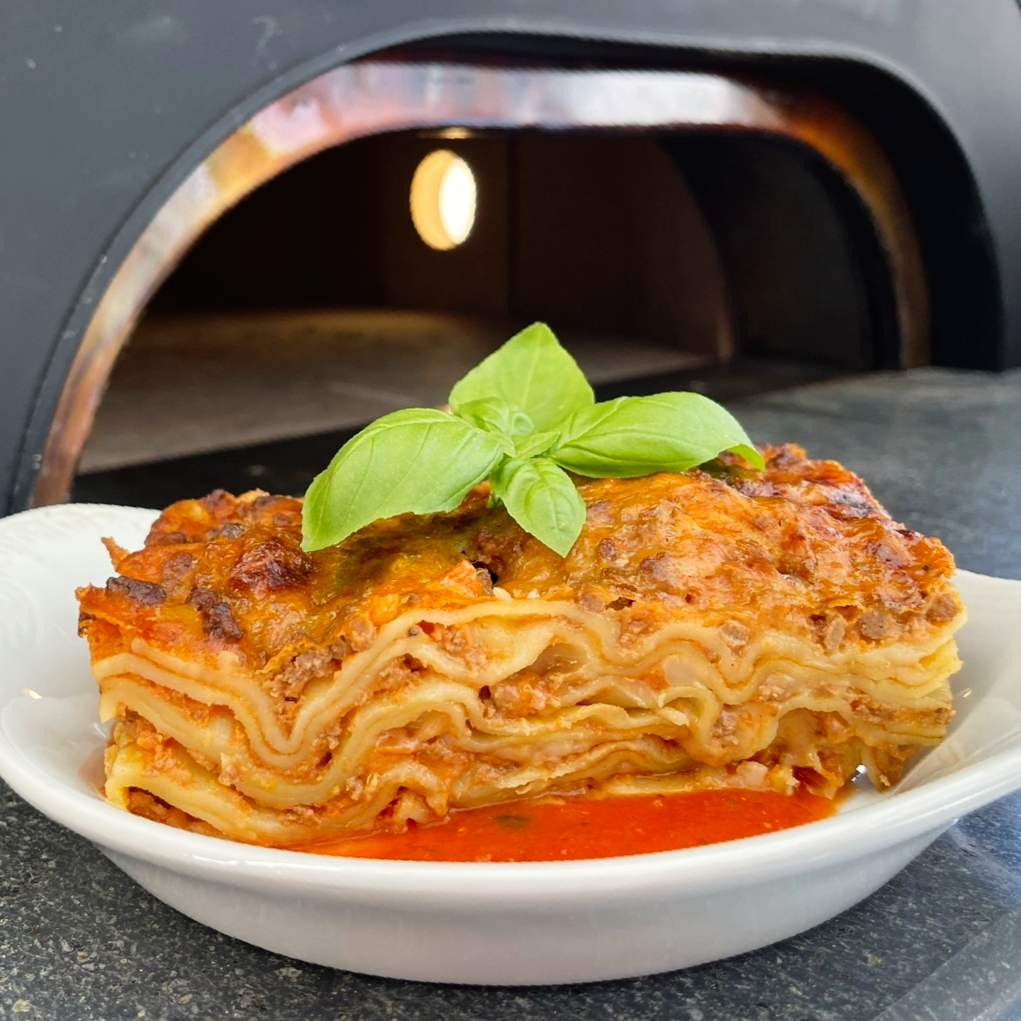Lasagna in a white bowl, topped with basil, in front of a pizza oven.