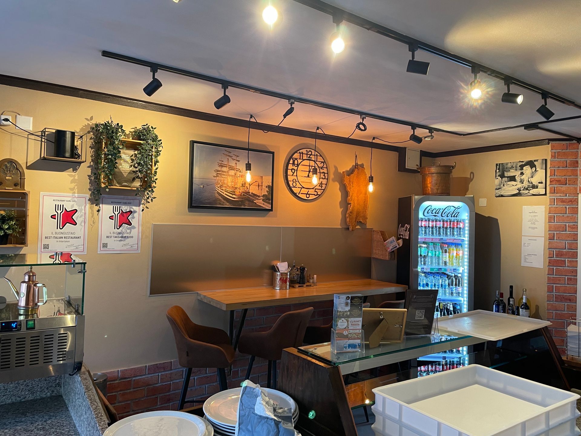 Innenansicht eines Cafés mit Tischen, Stühlen, Getränkekühler, Wandbildern und Schienenbeleuchtung.
