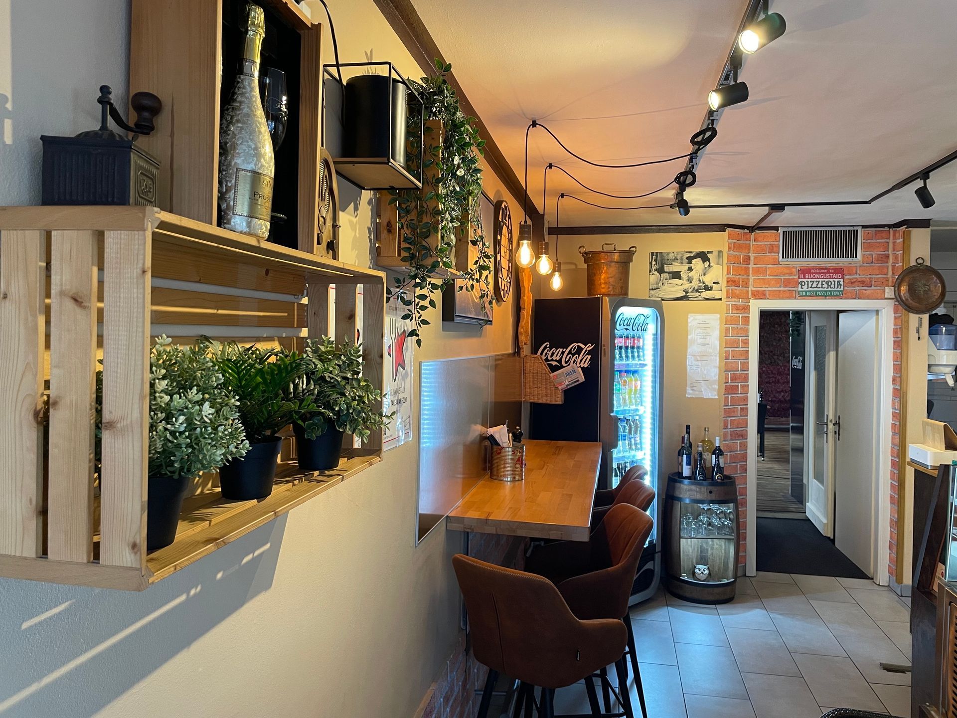 Innenansicht einer Bar mit als Regale dienenden Holzkisten. Pflanzen, eine Bartheke mit Stühlen und ein Kühlschrank.