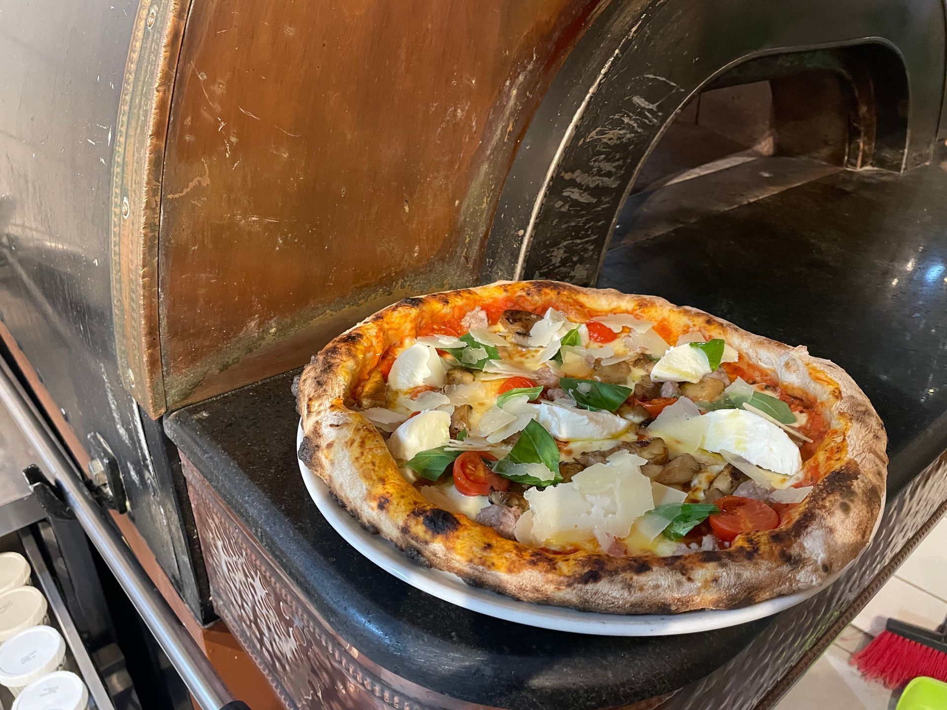 Pizza auf einem Teller neben einem Steinofen, belegt mit Käse, Tomaten und Basilikum.