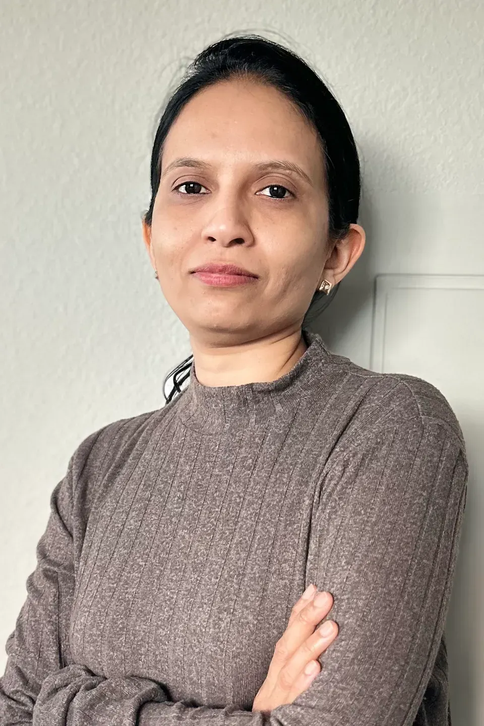 A woman in a brown sweater is standing with her arms crossed.