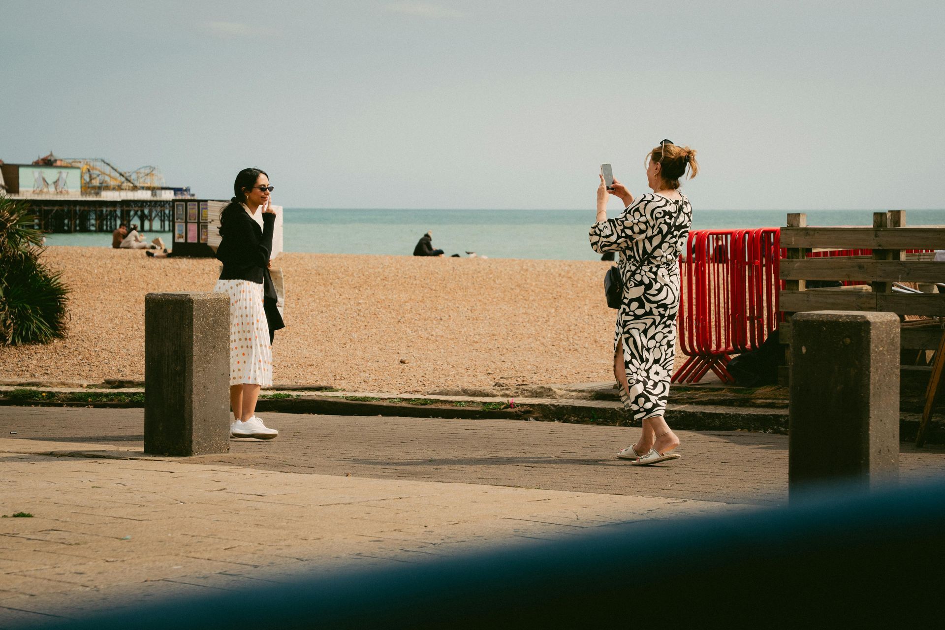 Two people stand on a paved path overlooking a beach; one poses while the other captures a photo with a smartphone.