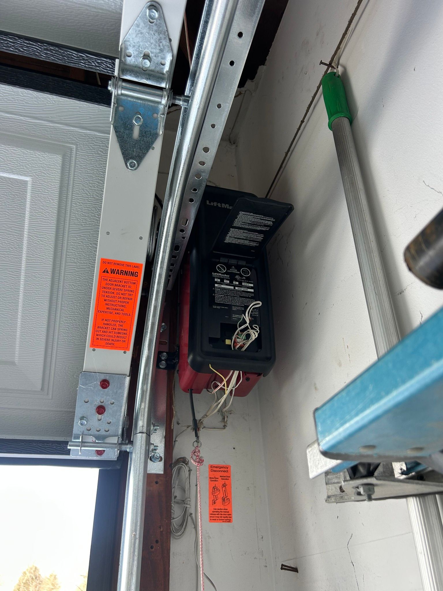 Garage door opener motor mounted on ceiling rail with wiring and orange warning labels