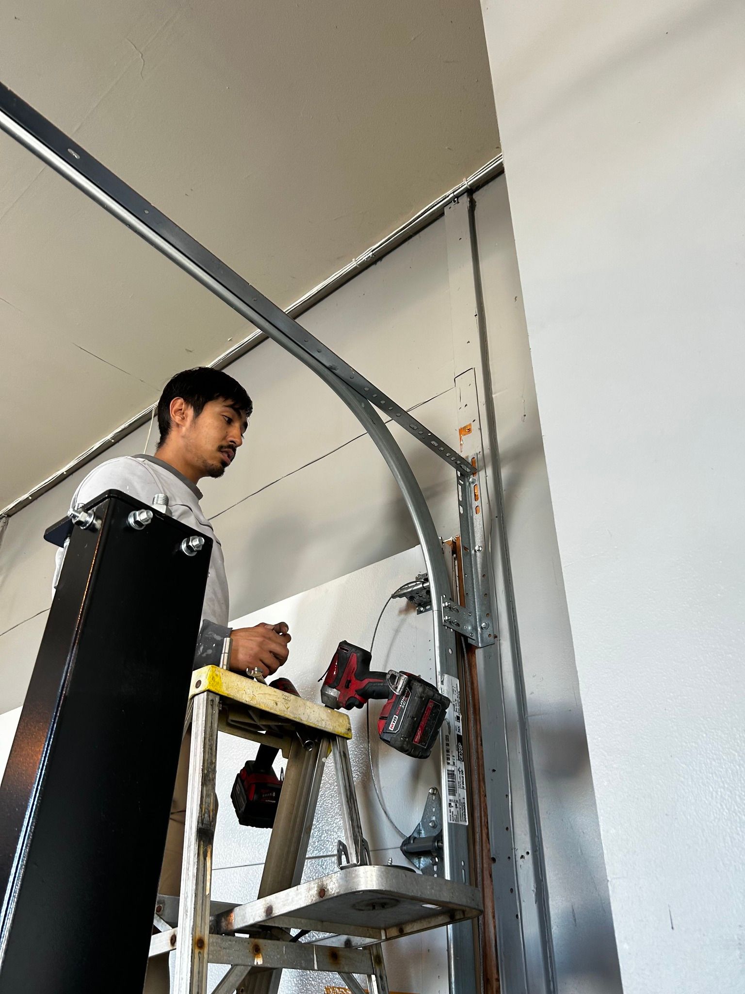 Man on a ladder working on a ceiling-mounted metal frame in a garage or workshop