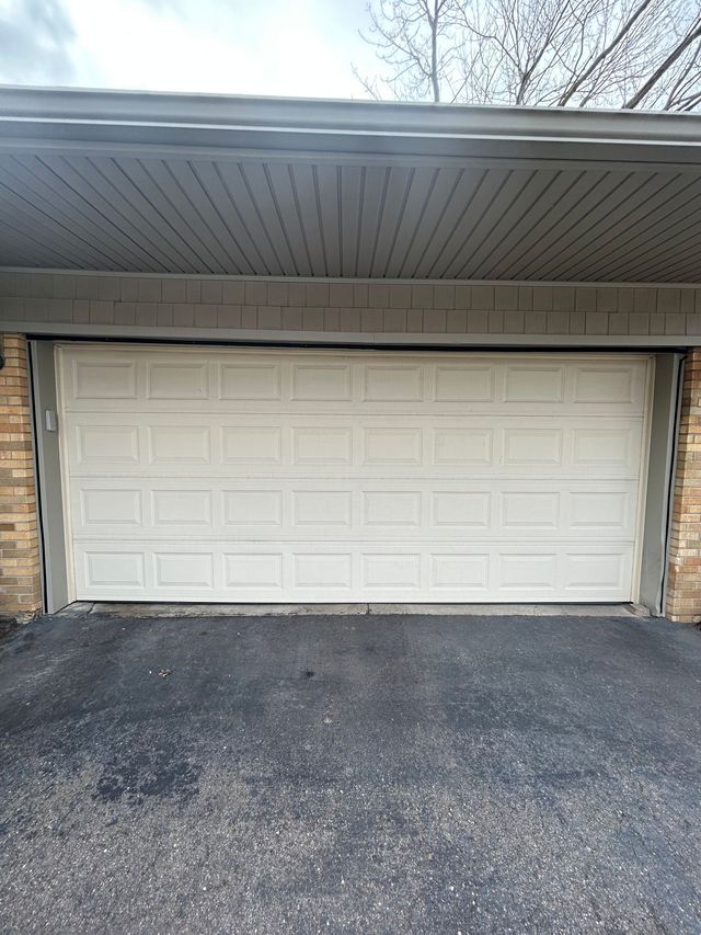 White garage door beneath a covered driveway with brick siding and a black asphalt driveway