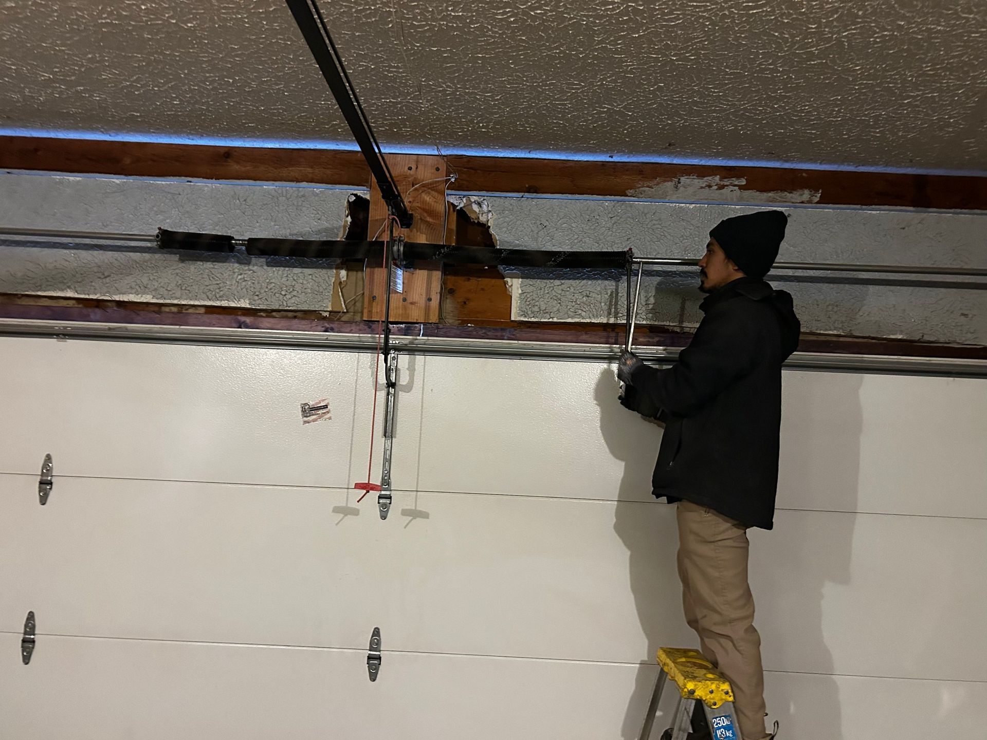 Worker standing on a stool adjusting a garage door opener rail near the ceiling.