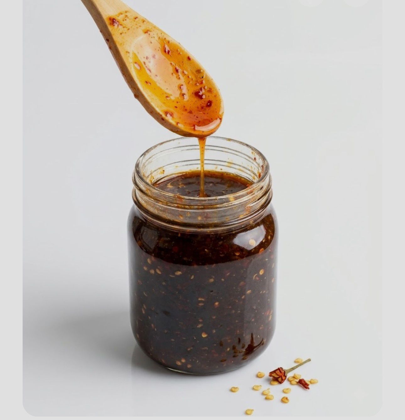 Jar of dark chili sauce with a wooden spoon dripping sauce above it, on a light background