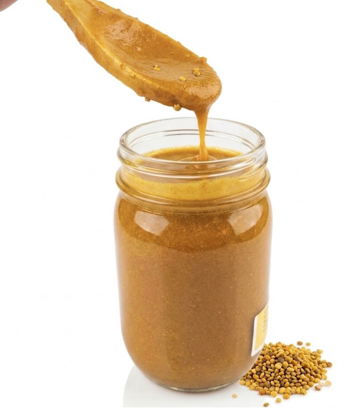 Spoon dripping golden mustard into a glass jar, with mustard seeds beside it.