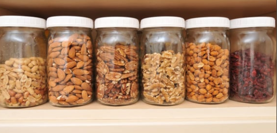 Six glass jars of assorted nuts and dried fruit lined up on a pantry shelf