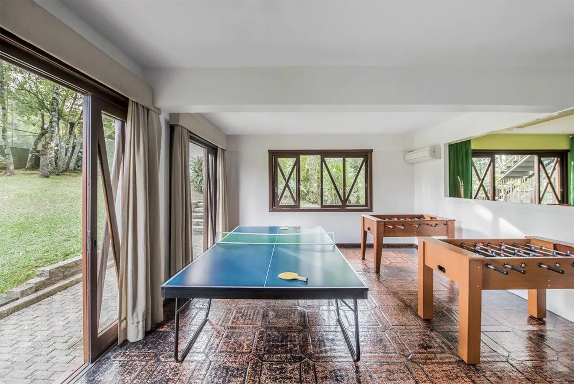 Sala de jogos ao ar livre com mesas de pingue-pongue e pebolim em um pátio coberto com janelas abertas.