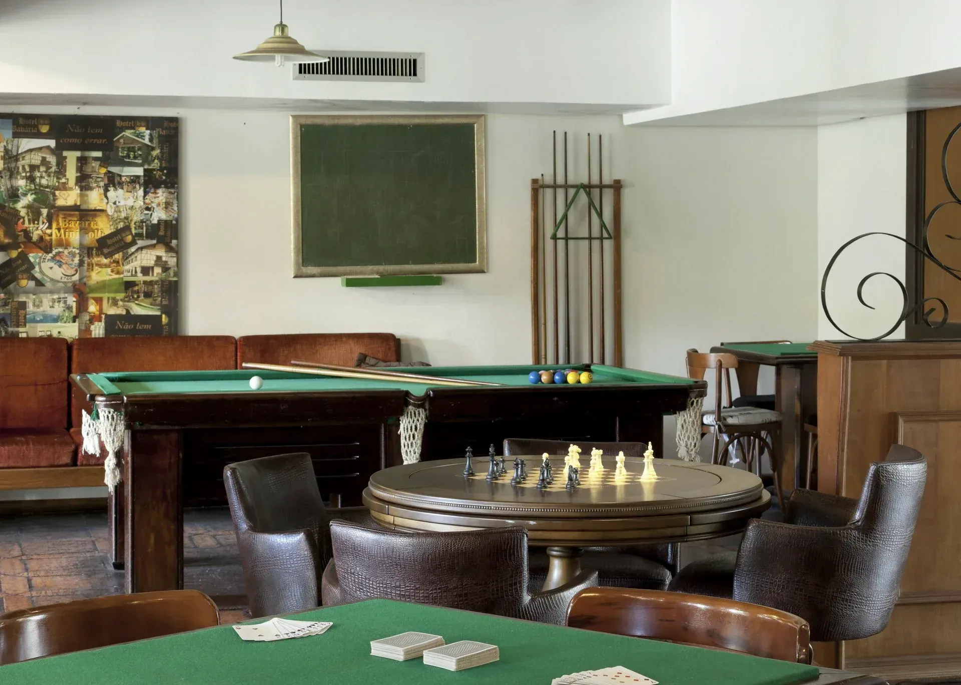 Sala de jogos com mesas de bilhar, tabuleiro de xadrez, cadeiras de couro e um quadro-negro na parede.