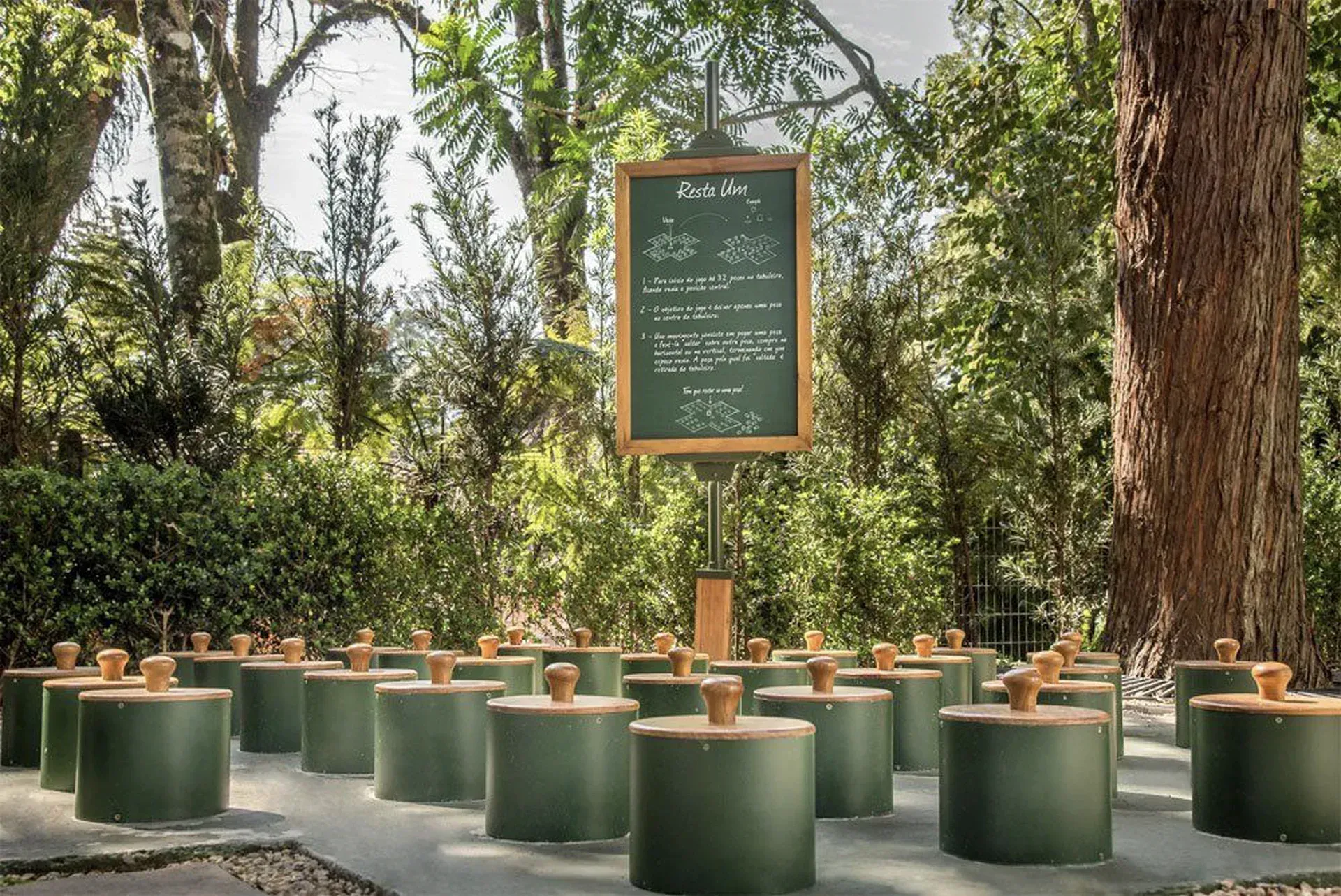 Memorial ao ar livre com bancos cilíndricos verdes em torno de um quadro-negro central, em um ambiente arborizado.