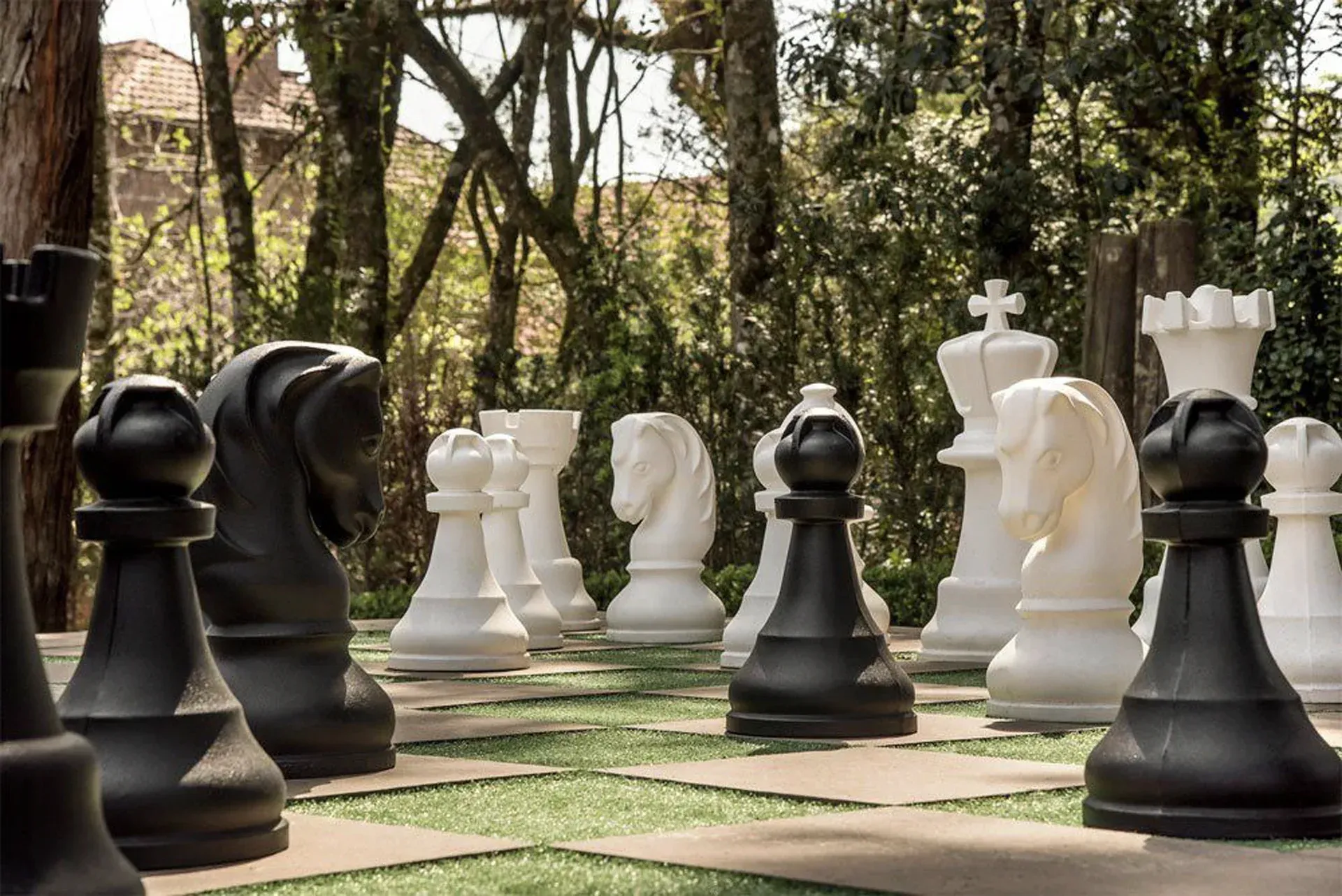 Grande jogo de xadrez ao ar livre com peças pretas e brancas de tamanho grande em um tabuleiro estilo parque entre árvores.