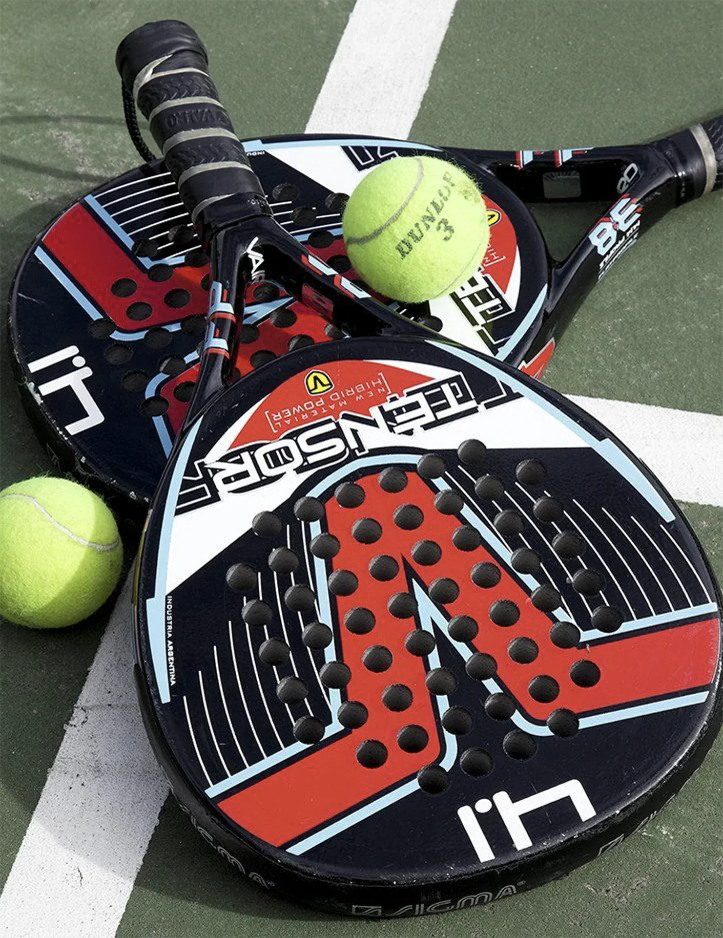 Duas raquetes de pickleball pretas e vermelhas com uma bola amarela em uma linha da quadra.