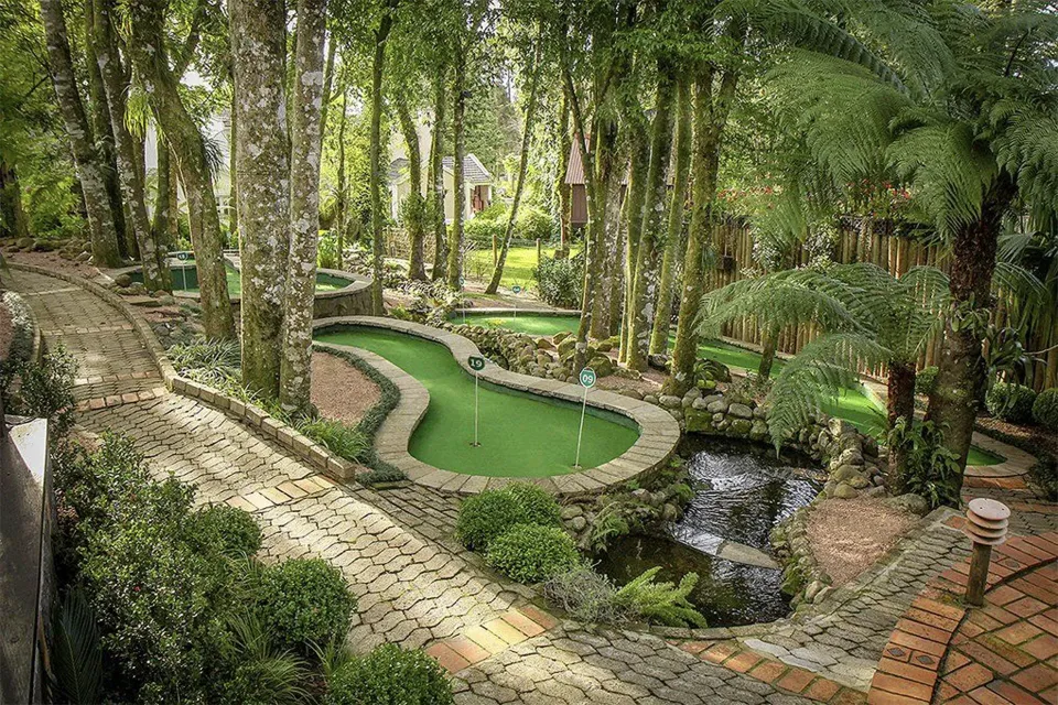 Jardim exuberante com um pequeno lago verde, caminhos de pedra e densas árvores tropicais.