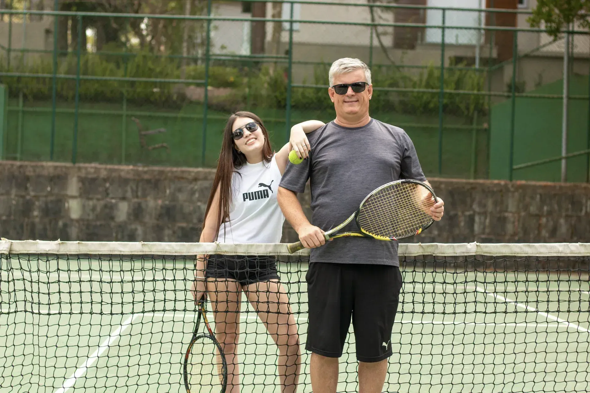 Duas pessoas posando em uma quadra de tênis, segurando raquetes e sorrindo atrás da rede.
