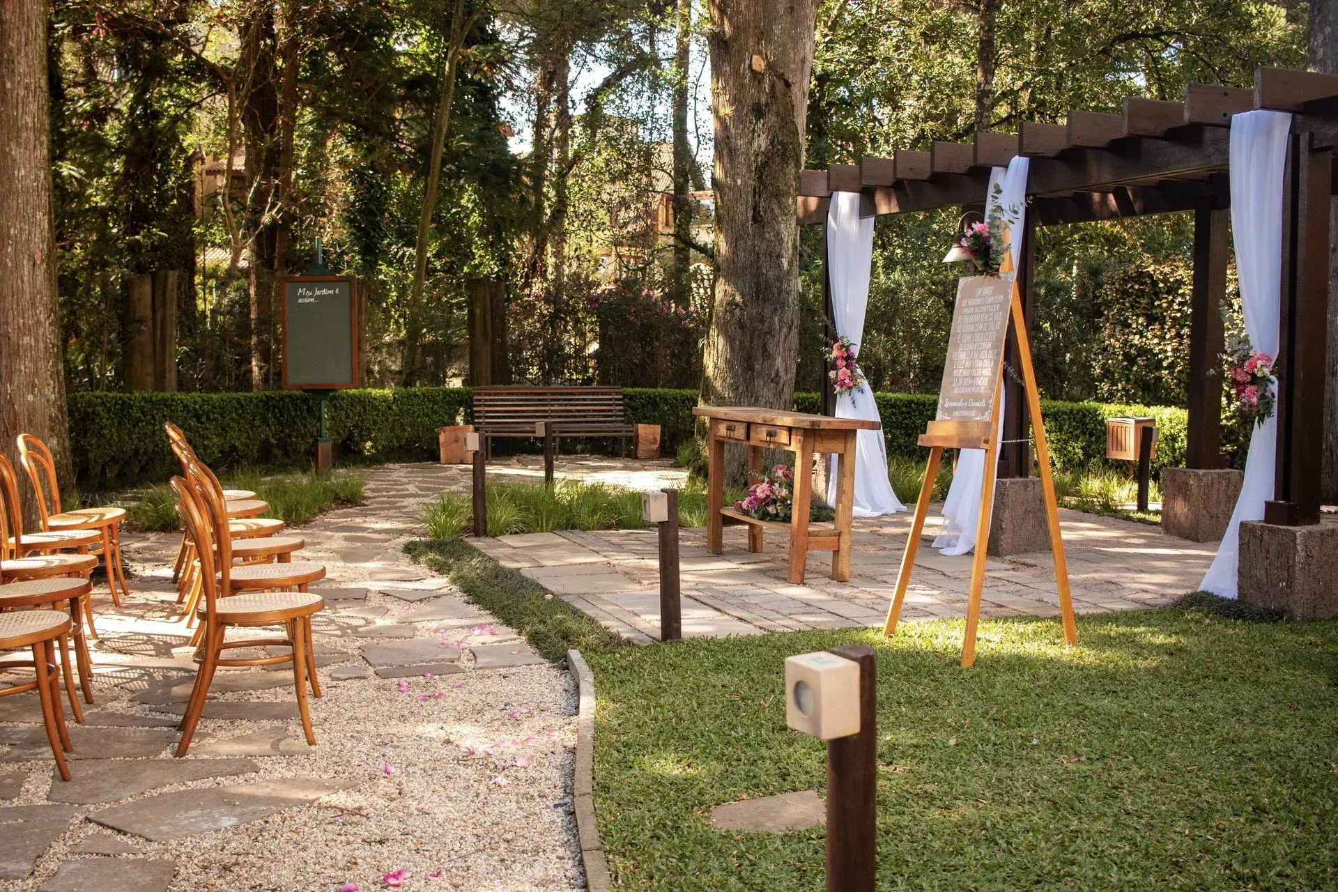 Evento ao ar livre em jardim, com cadeiras de madeira, cavaletes e um dossel sob as árvores.